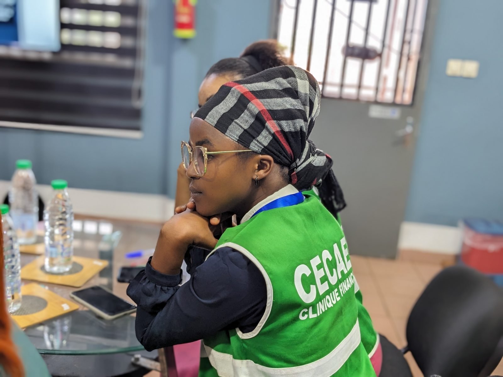De profil, une participante portant des lunettes et un gilet vert « CECAEC Clinique financière » suit la séance, le menton posé sur les mains.