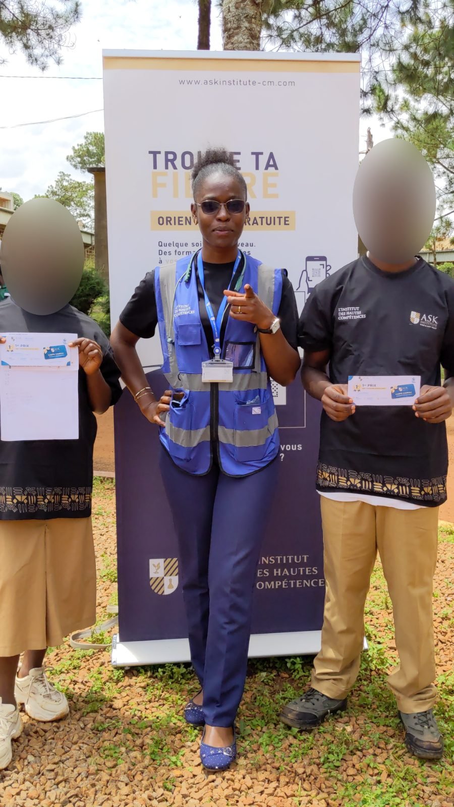 Une membre de l’équipe de ASK Institute, en gilet de l’Institut, pose entre les deux lauréats qui tiennent leur carte cadeau « 1er prix », devant le kakémono « Trouve ta spécialité ».