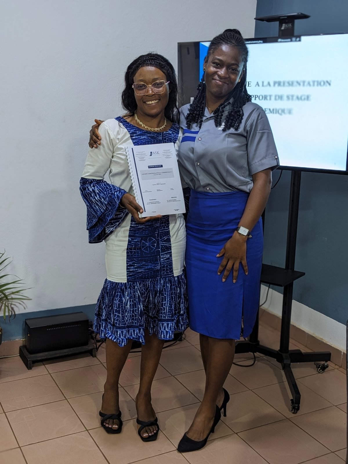 L’apprenante, rapport de stage en main, pose avec une membre de l’équipe de ASK Institute en uniforme de l’Institut, qui la tient par l’épaule.