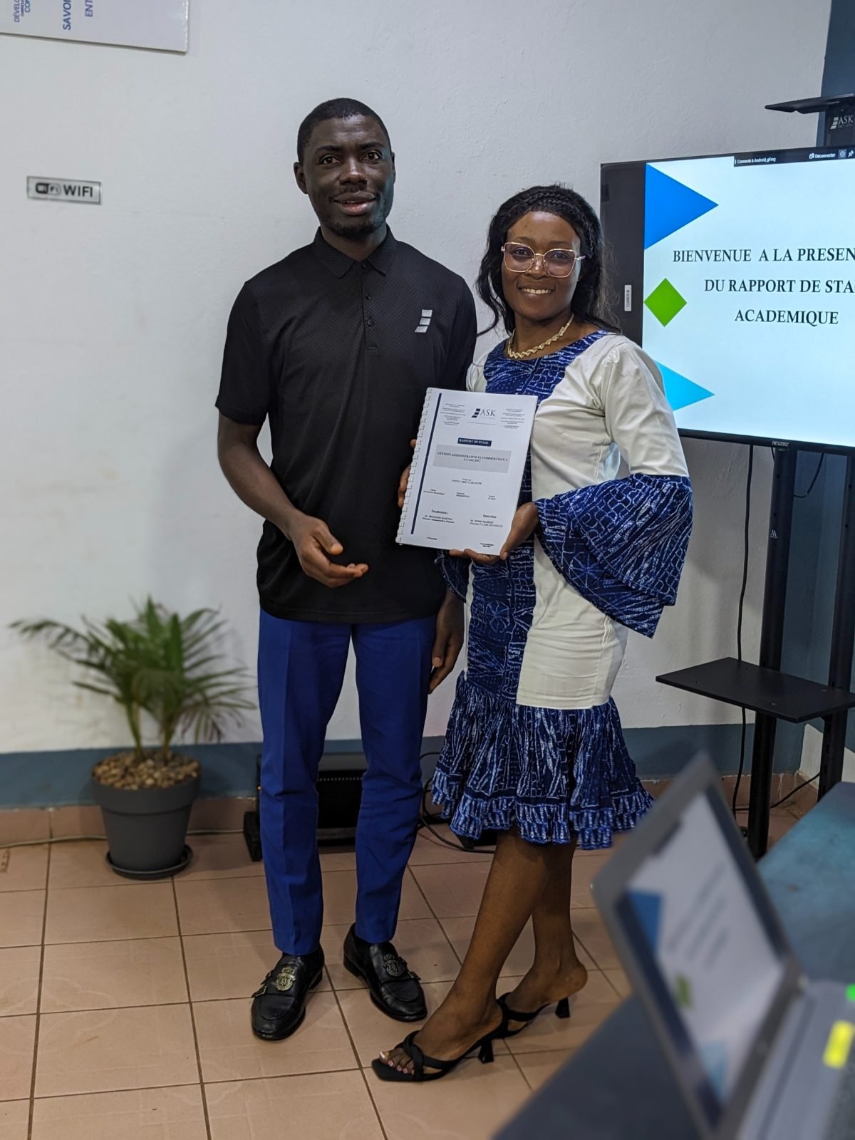 L’apprenante, son rapport de stage relié en main, pose aux côtés d’un membre du jury devant l’écran « Bienvenue à la présentation du rapport de stage académique ».